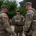 Three U.S. Army soldiers in Operational Camouflage Pattern (OCP) uniforms are engaged in a conversation outdoors, likely during a security or patrol mission. All three are wearing body armor/plate carriers and tactical gear. The setting appears to be an urban or institutional environment with trees and a modern building visible in the background.