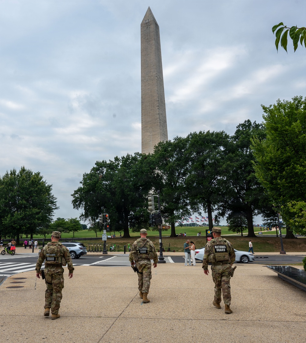 Three National Guard soldiers in camouflage uniforms are walking toward the Washington Monument on the National Mall in Washington, D.C. The soldiers are viewed from behind, with 'NATIONAL GUARD' patches clearly visible on their body armor. The scene shows a typical day on the Mall with civilian visitors, trees in full summer foliage, and American flags visible near the monument.