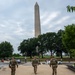 Three National Guard soldiers in camouflage uniforms are walking toward the Washington Monument on the National Mall in Washington, D.C. The soldiers are viewed from behind, with 'NATIONAL GUARD' patches clearly visible on their body armor. The scene shows a typical day on the Mall with civilian visitors, trees in full summer foliage, and American flags visible near the monument.