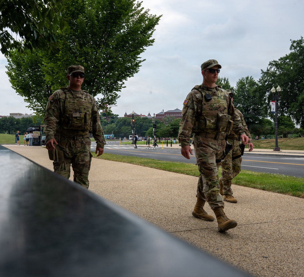 Two U.S. Army soldiers in Operational Camouflage Pattern (OCP) uniforms are conducting a foot patrol along a sidewalk in what appears to be Washington, D.C., based on the National Mall-area architecture and landscape visible in the background. Both soldiers are equipped with tactical gear, sidearms, and wear sunglasses. The setting suggests a security or ceremonial patrol mission in a public area.