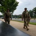Two U.S. Army soldiers in Operational Camouflage Pattern (OCP) uniforms are conducting a foot patrol along a sidewalk in what appears to be Washington, D.C., based on the National Mall-area architecture and landscape visible in the background. Both soldiers are equipped with tactical gear, sidearms, and wear sunglasses. The setting suggests a security or ceremonial patrol mission in a public area.