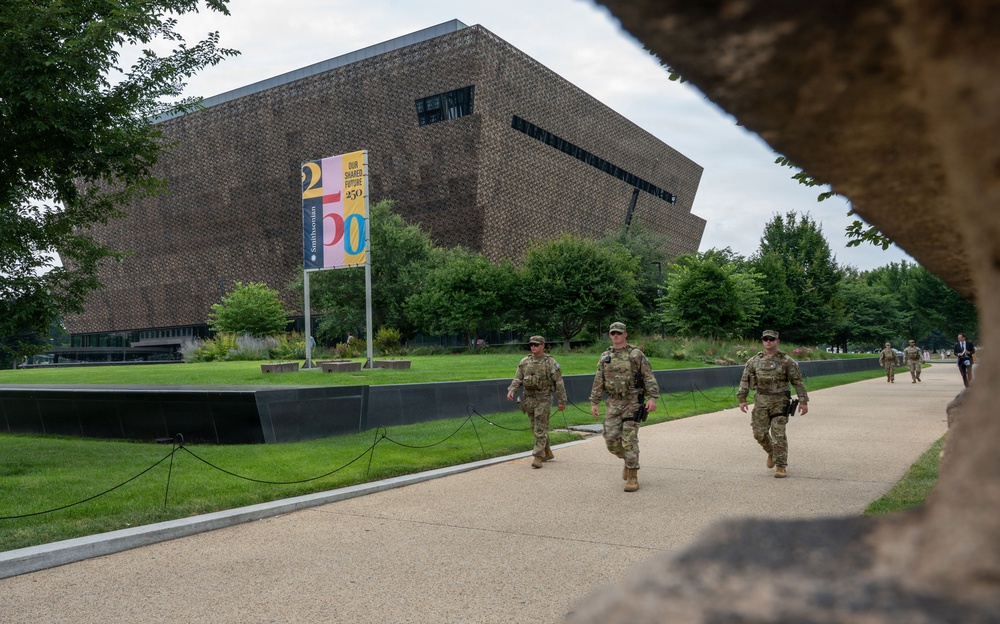 Military or National Guard personnel in camouflage uniforms are patrolling a walkway near the Smithsonian National Museum of African American History and Culture (NMAAHC) on the National Mall in Washington, D.C. A colorful Smithsonian banner reading '250' and 'Our Shared Future' is visible near the museum's distinctive bronze-latticed exterior. The scene suggests a period of heightened security presence at a major national institution.