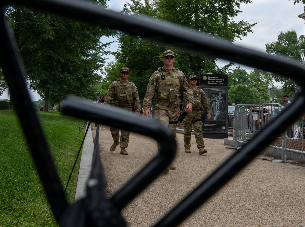 Three military personnel in camouflage uniforms are walking along a paved path near the Smithsonian National Museum of African American History and Culture in Washington, D.C. The image is photographed through black metal security fencing in the foreground, creating a framed perspective. The scene suggests a security or patrol presence near a major national institution.
