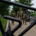 Three military personnel in camouflage uniforms are walking along a paved path near the Smithsonian National Museum of African American History and Culture in Washington, D.C. The image is photographed through black metal security fencing in the foreground, creating a framed perspective. The scene suggests a security or patrol presence near a major national institution.