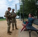 Two U.S. Army soldiers in OCP (Operational Camouflage Pattern) uniforms are conversing with a civilian woman seated on a park bench on the National Mall in Washington, D.C. The Washington Monument is clearly visible in the background. The interaction appears casual and friendly, suggesting a community engagement or public assistance role.