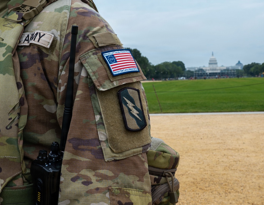 A close-up photograph of a U.S. Army soldier in Operational Camouflage Pattern (OCP) uniform, standing on the National Mall in Washington D.C. with the U.S. Capitol building visible in the background. The soldier's sleeve displays a District of Columbia National Guard patch with an American flag design and a lightning bolt unit insignia patch. The soldier is equipped with a radio/communications device on their vest.
