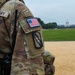 A close-up photograph of a U.S. Army soldier in Operational Camouflage Pattern (OCP) uniform, standing on the National Mall in Washington D.C. with the U.S. Capitol building visible in the background. The soldier's sleeve displays a District of Columbia National Guard patch with an American flag design and a lightning bolt unit insignia patch. The soldier is equipped with a radio/communications device on their vest.
