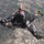 A skydiver is captured mid-freefall from a top-down perspective, wearing a black patterned jumpsuit and helmet with a mounted camera. The urban and agricultural landscape below suggests a significant altitude, likely several thousand feet. One leg appears to end in a prosthetic limb with a green athletic shoe attached.