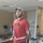 A bearded adult male is undergoing physical rehabilitation therapy in a clinical setting, standing between parallel bars with a gait belt around his waist. His left leg is fitted with a black orthopedic walking boot, while his right leg has an external fixator device (circular/ring fixator) attached from the knee area down to the foot, which is wrapped in a bandage. A wheelchair is visible behind him, and a therapist's hand is partially visible at the left edge of the frame.