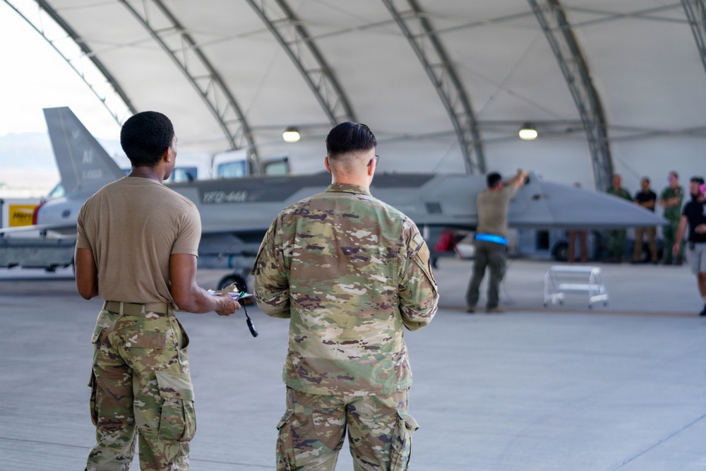 Two military personnel in OCP (Operational Camouflage Pattern) uniforms stand with their backs to the camera, observing activity around a military jet aircraft inside a large hangar structure. The aircraft, which appears to bear markings consistent with a modern fighter jet, is being attended to by ground crew personnel. The setting appears to be a military air base with an arched hangar shelter.