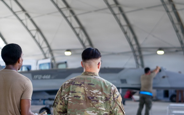 Two military personnel in OCP (Operational Camouflage Pattern) uniforms stand with their backs to the camera, observing activity around a military jet aircraft inside a large hangar structure. The aircraft, which appears to bear markings consistent with a modern fighter jet, is being attended to by ground crew personnel. The setting appears to be a military air base with an arched hangar shelter.