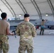 Two military personnel in OCP (Operational Camouflage Pattern) uniforms stand with their backs to the camera, observing activity around a military jet aircraft inside a large hangar structure. The aircraft, which appears to bear markings consistent with a modern fighter jet, is being attended to by ground crew personnel. The setting appears to be a military air base with an arched hangar shelter.