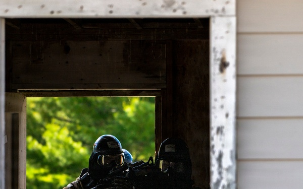 Two heavily equipped tactical operators are visible through a weathered window frame of a structure, appearing to conduct a building clearance or entry drill. Both personnel are wearing full tactical gear including ballistic helmets, face protection, body armor, and are carrying assault rifles. The setting and controlled nature of the scene suggest a military or law enforcement training exercise.