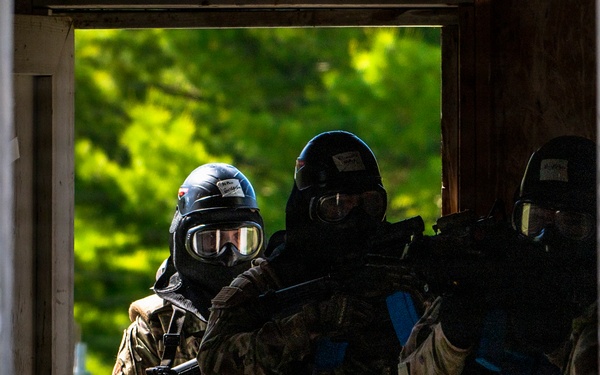 Three heavily equipped tactical operators are visible through a window frame of a dilapidated building, wearing full protective gear including helmets, gas masks, and body armor. They are carrying firearms and appear to be conducting a military or law enforcement training exercise. The setting suggests a controlled urban operations or close-quarters battle (CQB) training environment.