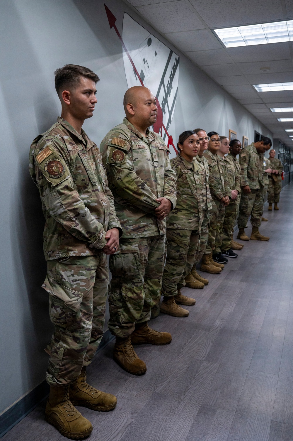 A group of approximately 10-12 military personnel in OCP (Operational Camouflage Pattern) uniforms are standing in a line along a corridor wall. They appear to be at attention or in a relaxed standing position, likely during a formal or semi-formal military event. The setting appears to be an indoor military facility with decorative wall art featuring red and black geometric designs.