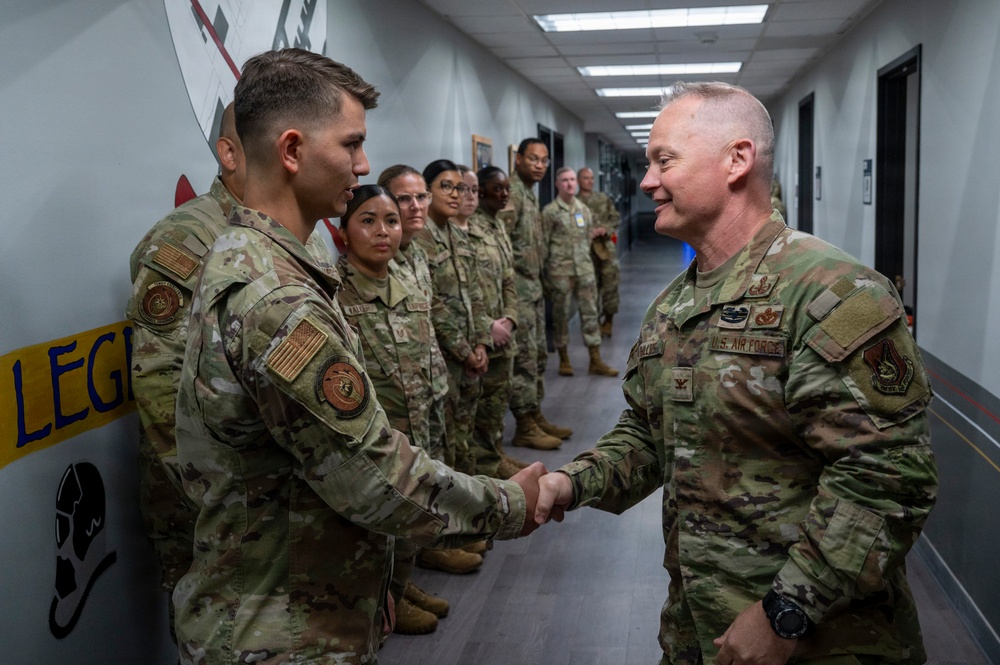 A senior U.S. Air Force officer, identifiable by colonel rank insignia and 'U.S. AIR FORCE' tape, is shaking hands with a junior airman in a military corridor. A formation of uniformed personnel in OCP (Operational Camouflage Pattern) uniforms stands lined up along the wall in the background. The setting appears to be an official visit or recognition event inside a military facility.