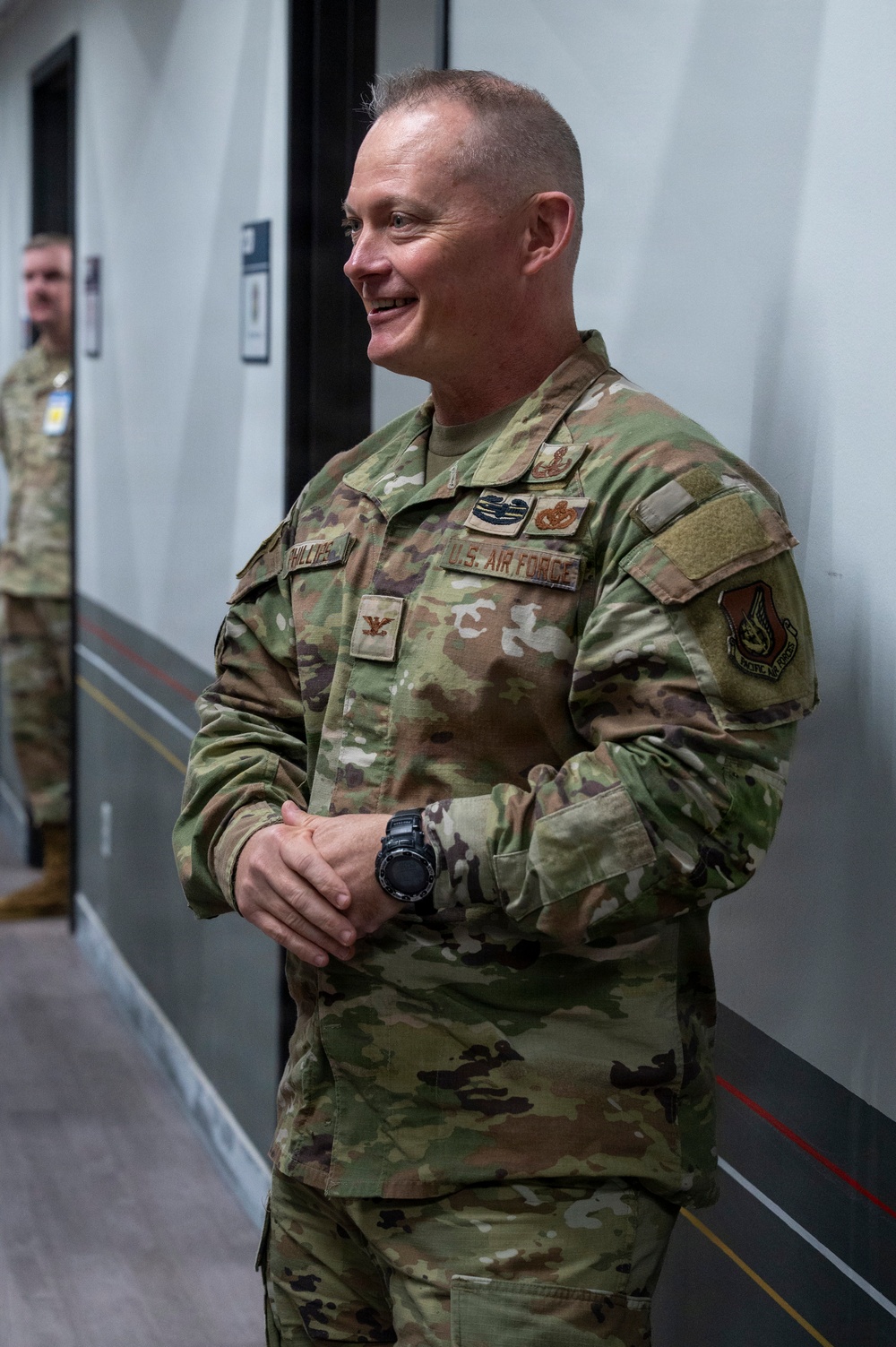 A U.S. Air Force officer in OCP (Operational Camouflage Pattern) uniform is standing in an indoor corridor, smiling. The individual displays rank insignia and branch tape visible on the uniform. A second uniformed service member is partially visible in the background.