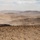 A vast arid desert landscape photographed from an elevated vantage point, showing rolling hills, sparse desert scrub vegetation, and a winding dirt road in the valley below. A small group of people is visible as silhouettes along a ridgeline in the middle distance, appearing very small against the expansive terrain. The scene is hazy with atmospheric dust, suggesting hot, dry conditions.