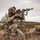 A soldier in full combat gear is kneeling in a desert/arid terrain while aiming an assault rifle. The individual is equipped with multicam camouflage uniform, tactical vest, helmet with camouflage cover, and optical sighting equipment. The setting appears to be a training exercise or field operation in a rocky, scrub-brush desert environment.