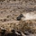 A tracked military armored fighting vehicle is moving across an arid desert terrain, kicking up a significant dust cloud behind it. The landscape is characterized by sparse desert scrub vegetation, rocky soil, and flat open terrain typical of a desert training environment. The vehicle appears to be traveling at speed along a dirt track.