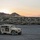 A tan/desert-camouflage military HMMWV (Humvee) is parked on a flat, arid desert terrain at sunset. The vehicle bears the number '624' and unit insignia on its door, with a radio antenna extended from the rear. The scene is set against a backdrop of low desert mountains with a warm orange and pink sunset sky.