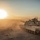 A main battle tank, consistent with an M1 Abrams, is positioned in an arid desert environment during sunset or sunrise. At least one crew member is visible in the open hatch atop the turret. The scene is bathed in warm golden light with dust haze and distant mountain ridgelines visible on the horizon.