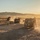 A convoy of military tactical vehicles is moving across an arid desert terrain, kicking up dust trails in golden sunset lighting. At least three vehicles are visible traveling in a loose column formation across flat, scrub-covered desert ground. Mountain ranges are visible in the background under a hazy, sun-lit sky.