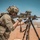 A heavily equipped military sniper is positioned on a rooftop in a desert environment, aiming a large precision rifle mounted on a bipod tripod. The soldier is wearing full tactical gear including a camouflage uniform, plate carrier vest, and helmet with attachments. The setting appears to be a training range or operational environment in an arid, desert region.