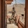 Two soldiers in full combat gear are positioned on a rooftop balcony of a tan/adobe-style building in an arid desert environment. One soldier is standing and using binoculars while the other is crouching and operating what appears to be a tripod-mounted optical or communications device. A rifle is leaned against the balcony railing nearby.