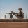 A military soldier in full combat gear stands on a rooftop position behind a low concrete block wall. The soldier is holding a large sniper rifle mounted on a tripod, appearing to be in an overwatch or security position. The arid, hazy environment suggests a desert operational theater.