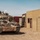 A desert-camouflaged Bradley Fighting Vehicle (IFV) is positioned in what appears to be a military training facility or mock urban environment. The vehicle is equipped with camouflage netting, a coil of concertina wire, and various electronic systems on its turret. The setting features simulated building structures and shipping containers consistent with a combat training center.