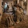 Two armed soldiers in desert camouflage uniforms are positioned inside a damaged building with ornate carpeted floors. The foreground soldier is crouching against a wall while holding a rifle and communicating via radio or phone. The setting appears to be an interior clearing or security operation within a deteriorated structure.