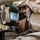 A close-up photograph of a military personnel looking through an optical sighting device, likely a weapon or artillery sight. The soldier is wearing full tactical gear including a helmet with cover, ballistic eyewear, and communications headset. The image is a high-quality, sharp photograph taken in what appears to be an indoor or sheltered military environment.