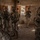 A group of military personnel in full combat gear are gathered inside a cinder block structure, appearing to receive a briefing or instructions from one soldier who is gesturing. The setting appears to be a desert training environment, likely a military exercise at a facility such as the National Training Center (NTC) in California. The soldiers are equipped with rifles, tactical vests, helmets with camouflage netting, and heavy rucksacks.