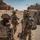 A group of military personnel in full combat gear are advancing on foot alongside an armored vehicle in a desert training environment. The soldiers are equipped with various weapons and tactical equipment, moving toward a simulated urban structure. The setting appears to be a military training exercise, likely at a facility such as the National Training Center (NTC) at Fort Irwin, California.
