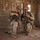 Two armed military personnel in full combat gear are crouched inside a cinder block structure with a sandy floor. They appear to be reviewing information on a small device while holding assault rifles. The setting suggests a training exercise or operational environment in an arid region.