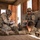 Three military personnel in U.S. Army combat uniforms are inside an unfinished concrete block structure, likely during a training exercise. Two soldiers are kneeling/crouching and writing on notepads or tablets while a third soldier lies prone on the sandy floor. The setting appears to be a military training facility, possibly in a desert environment.