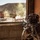 A heavily equipped soldier in full camouflage gear, including a ghillie-style helmet cover, is positioned at a window inside a building overlooking a desert training environment. Smoke or dust clouds are visible rising from structures in the background, suggesting an active exercise or simulated engagement. The setting appears to be a military urban warfare training facility in an arid, desert landscape.