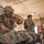 Three soldiers in U.S. Army MultiCam (OCP) uniforms are performing tactical combat casualty care (TCCC) on a simulated or actual casualty lying on a field stretcher inside a concrete block structure. The soldier on the left is applying or tightening a tourniquet or bandage, while the female soldier on the right appears to be documenting or administering additional treatment. The setting and equipment suggest a training exercise or forward aid station scenario.