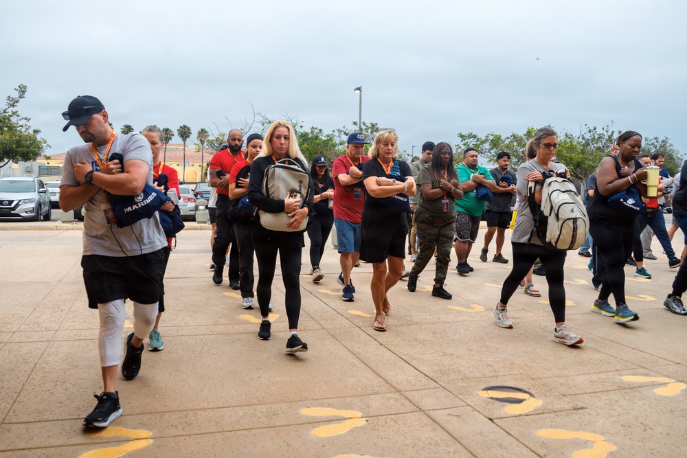 A large group of civilians are marching in formation across a paved outdoor area marked with yellow footprint symbols, characteristic of a Marine Corps recruit depot experience. Participants are carrying blue 'Marines' branded tote bags and backpacks while walking in rows with arms crossed or held close to their bodies. The setting appears to be a military base, likely part of a civilian orientation or 'Educator Workshop' program.