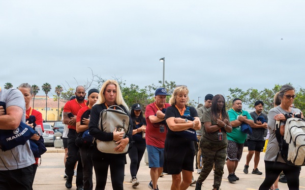 A large group of civilians are marching in formation across a paved outdoor area marked with yellow footprint symbols, characteristic of a Marine Corps recruit depot experience. Participants are carrying blue 'Marines' branded tote bags and backpacks while walking in rows with arms crossed or held close to their bodies. The setting appears to be a military base, likely part of a civilian orientation or 'Educator Workshop' program.