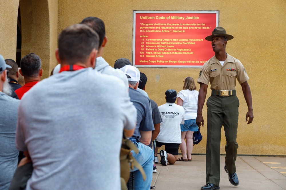 A U.S. Marine Corps drill instructor in uniform addresses a group of civilians gathered near a wall displaying a 'Uniform Code of Military Justice' sign. The setting appears to be a military base, likely during a family day, recruit graduation event, or public orientation. The drill instructor is speaking authoritatively while the group listens attentively.