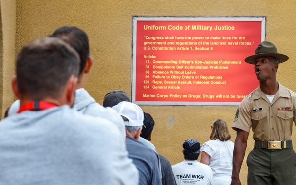 A U.S. Marine Corps drill instructor in uniform addresses a group of civilians gathered near a wall displaying a 'Uniform Code of Military Justice' sign. The setting appears to be a military base, likely during a family day, recruit graduation event, or public orientation. The drill instructor is speaking authoritatively while the group listens attentively.