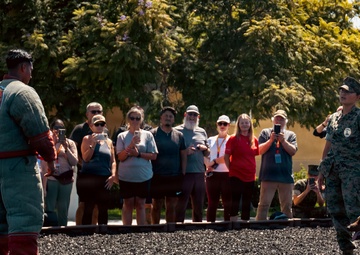 A military working dog demonstration is taking place outdoors, with a uniformed handler and a dog in a low crouch position facing a person wearing a padded bite suit. A crowd of civilian observers, many photographing with phones, watches from behind a boundary line. The setting appears to be a military installation, indicated by the camouflage uniform and institutional architecture.