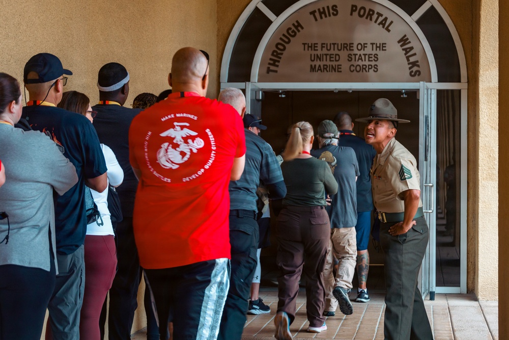 A group of civilians and a Marine Corps Drill Instructor are gathered at the entrance of a United States Marine Corps facility. The arched doorway bears the inscription 'Through This Portal Walks The Future Of The United States Marine Corps,' indicating this is likely a recruit processing or orientation event. A Drill Instructor in uniform appears to be directing or addressing the group as they enter the building.