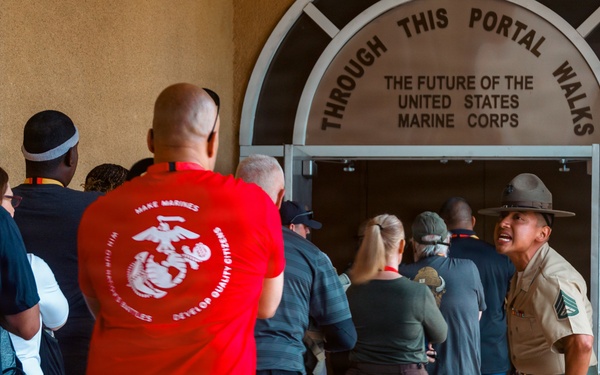 A group of civilians and a Marine Corps Drill Instructor are gathered at the entrance of a United States Marine Corps facility. The arched doorway bears the inscription 'Through This Portal Walks The Future Of The United States Marine Corps,' indicating this is likely a recruit processing or orientation event. A Drill Instructor in uniform appears to be directing or addressing the group as they enter the building.