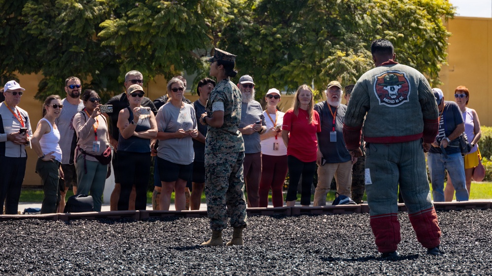 A military personnel in camouflage uniform addresses a civilian group at what appears to be a Marine Corps base, likely during a public tour or family day event. A second individual wearing a padded training suit with an 'MCRD K-9' patch stands nearby, suggesting a military working dog demonstration. The civilians are gathered outdoors on a gravel surface, many wearing visitor badges and recording with phones.