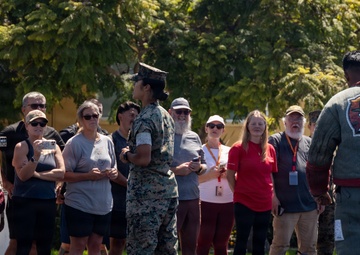 A military personnel in camouflage uniform addresses a civilian group at what appears to be a Marine Corps base, likely during a public tour or family day event. A second individual wearing a padded training suit with an 'MCRD K-9' patch stands nearby, suggesting a military working dog demonstration. The civilians are gathered outdoors on a gravel surface, many wearing visitor badges and recording with phones.