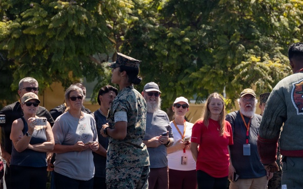 A military personnel in camouflage uniform addresses a civilian group at what appears to be a Marine Corps base, likely during a public tour or family day event. A second individual wearing a padded training suit with an 'MCRD K-9' patch stands nearby, suggesting a military working dog demonstration. The civilians are gathered outdoors on a gravel surface, many wearing visitor badges and recording with phones.