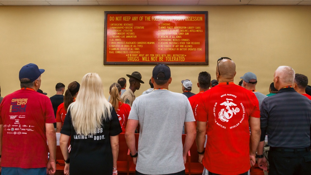 A group of civilians and at least one uniformed military drill instructor stand facing a large red warning sign listing prohibited items at what appears to be a U.S. Marine Corps recruit processing facility. The sign prominently states 'DO NOT KEEP ANY OF THE FOLLOWING IN YOUR POSSESSION' and ends with 'DRUGS WILL NOT BE TOLERATED.' This appears to be a recruit orientation or a civilian visitor tour event at a Marine Corps base.